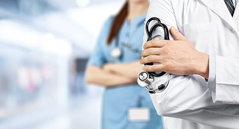 A doctor holding a stethoscope against his arm with a nurse with crossed arms in the background